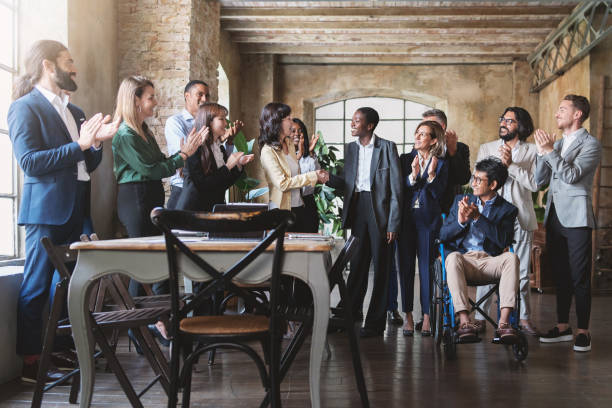 Diverse work team celebrates success in a modern rustic office. Middle-aged Japanese woman congratulates young African woman, as others applaud.