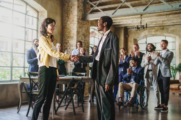 Multiethnic businesswomen shaking hands in front of their colleagues applauding in background - business lifestyle concept